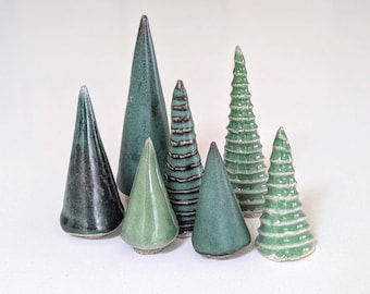 May include: A collection of seven ceramic tree figurines in various shades of green. The set includes smooth conical shapes in dark and light green, alongside textured pieces with horizontal ridges and layered, tiered designs.