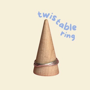 May include: A silver twistable ring on a wooden ring holder. The text "twistable ring" is written in blue above the ring.