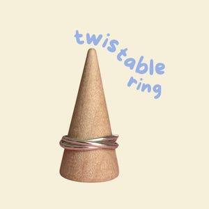 May include: A silver twistable ring on a wooden ring display stand. The text "twistable ring" is written in blue above the ring.