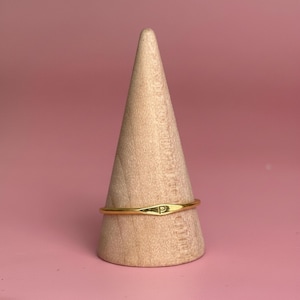 May include: A gold-colored ring with the letter "P" engraved on it, displayed on a wooden cone-shaped ring holder. The ring is thin and has a slightly curved design. The background is a solid pink color.