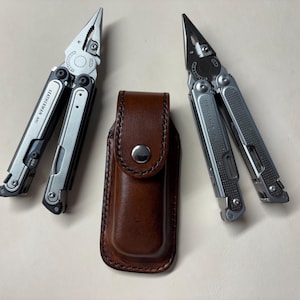 May include: Two silver and black multi-tools with the brand name "LEATHERMAN ARC" are displayed, flanking a brown leather sheath. The tools are open, showcasing their various components. The sheath is closed with a snap.