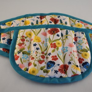 May include: A quilted fabric oven mitt with a white background featuring a colorful floral and strawberry print. The mitt is edged with solid teal fabric trim and includes matching teal fabric straps for hanging or securing the item during use.