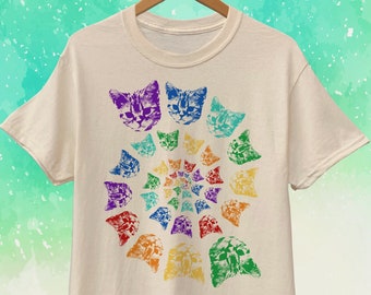 May include: Cream-colored t-shirt featuring a spiral design of colorful cat faces. The cat faces are in shades of purple, blue, green, yellow, orange, and red. The shirt is hanging on a wooden hanger.