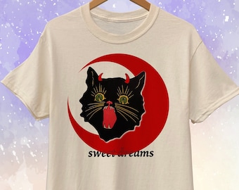 Op de afbeelding: Crèmekleurig T-shirt met een zwarte kat met rode hoorns en tong, tegen een rode halve maan. De tekst "sweet dreams" staat eronder. Het T-shirt hangt aan een houten kleerhanger.