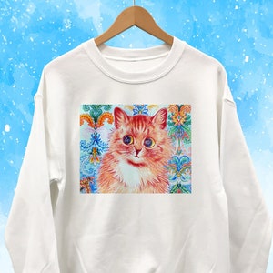 May include: White sweatshirt featuring a colorful cat portrait. The cat has orange fur, blue eyes, and a white chest. The background of the portrait is a decorative pattern with floral and geometric elements in shades of blue, orange, and green.