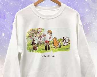 Może przedstawiać: Biała bluza z okrągłym dekoltem z kolorową ilustracją chłopca i postaci zwierząt na polu. Tekst "silly old bear" jest wydrukowany poniżej. Bluza wisi na drewnianym wieszaku.
