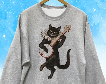 May include: A gray sweatshirt featuring a black cat playing a banjo. The cat wears a ruffled collar and has yellow eyes. The banjo is red and brown. The sweatshirt is hanging on a wooden hanger against a blue and white background.