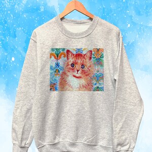 May include: A light gray sweatshirt featuring a colorful illustration of a cat with orange and white fur. The cat has large, expressive eyes and is set against a decorative background with floral and geometric patterns. The sweatshirt is hanging on a wooden hanger.