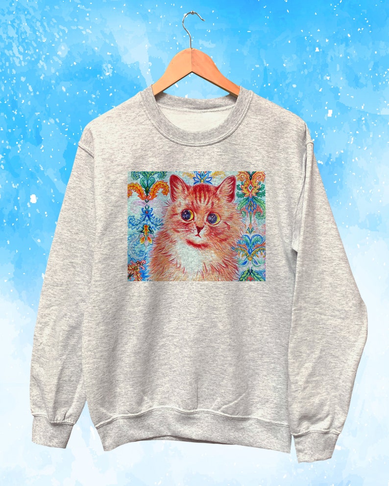 May include: A light gray sweatshirt featuring a colorful illustration of a cat with orange and white fur. The cat has large, expressive eyes and is set against a decorative background with floral and geometric patterns. The sweatshirt is hanging on a wooden hanger.