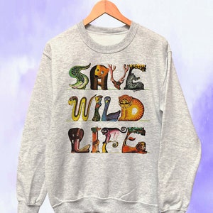 May include: A heather gray sweatshirt with the words "SAVE WILD LIFE" in colorful, animal-shaped letters. The design includes a crocodile, bear, deer, zebra, snake, bird, tiger, elephant, monkey, and lion. The sweatshirt is hanging on a wooden hanger.