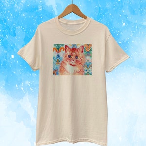 May include: Cream-colored t-shirt featuring a colorful cat portrait. The cat has orange fur, blue eyes, and a white chest. The background is a decorative pattern with floral and ornamental elements in shades of blue, green, and orange.