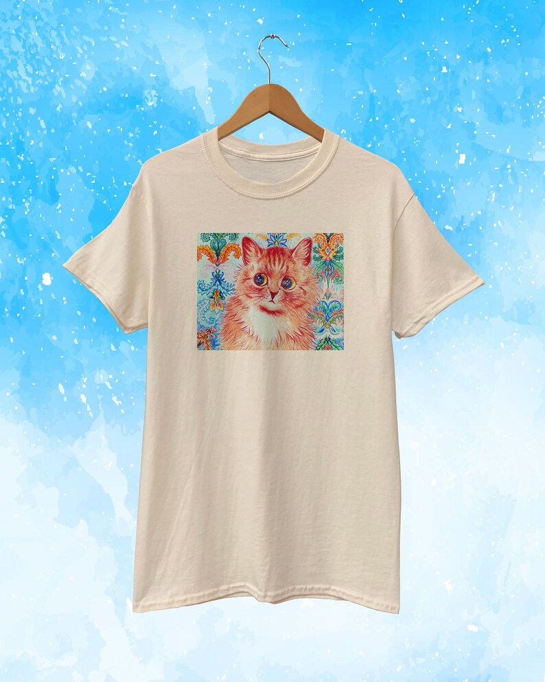 May include: Cream-colored t-shirt featuring a colorful cat portrait. The cat has orange fur, blue eyes, and a white chest. The background is a decorative pattern with floral and ornamental elements in shades of blue, green, and orange.