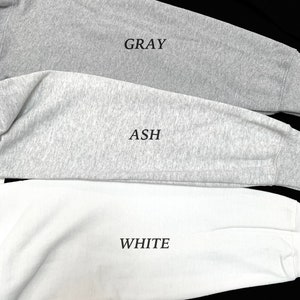 May include: Close-up of three long-sleeved shirts in gray, ash, and white. The word "GRAY" is printed on the gray shirt, "ASH" on the ash shirt, and "WHITE" on the white shirt. The shirts are made of a soft, comfortable material.