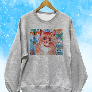 May include: A heather gray sweatshirt featuring a detailed illustration of a ginger cat with blue eyes. The cat is set against a backdrop of colorful floral and ornamental patterns. The sweatshirt is hanging on a wooden hanger.
