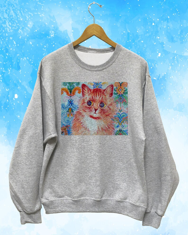 May include: A heather gray sweatshirt featuring a detailed illustration of a ginger cat with blue eyes. The cat is set against a backdrop of colorful floral and ornamental patterns. The sweatshirt is hanging on a wooden hanger.