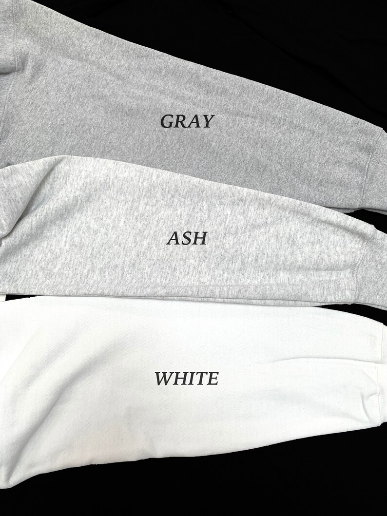 May include: Close-up of three long-sleeved shirts in gray, ash, and white. The word "GRAY" is printed on the gray shirt, "ASH" on the ash shirt, and "WHITE" on the white shirt. The shirts are made of a soft, textured fabric.