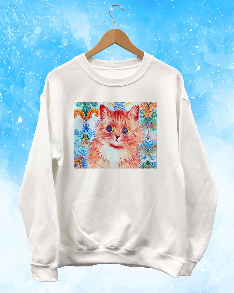 May include: White sweatshirt featuring a colorful cat portrait. The cat has orange fur, blue eyes, and a white chest. The background of the portrait is a decorative pattern with floral and geometric elements in shades of blue, orange, and green.