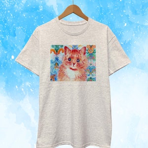 May include: A light gray t-shirt featuring a colorful cat portrait. The cat has orange fur, blue eyes, and is set against a decorative background with floral and ornamental patterns in shades of blue, orange, and green.