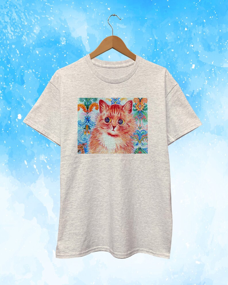 May include: A light gray t-shirt featuring a colorful cat portrait. The cat has orange fur, blue eyes, and is set against a decorative background with floral and ornamental patterns in shades of blue, orange, and green.
