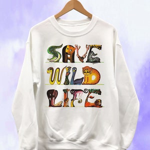 May include: White sweatshirt with the words "SAVE WILD LIFE" in colorful animal-shaped letters. The design includes a crocodile, bear, zebra, snake, bird, tiger, elephant, monkey, and lion. The sweatshirt is hanging on a wooden hanger.