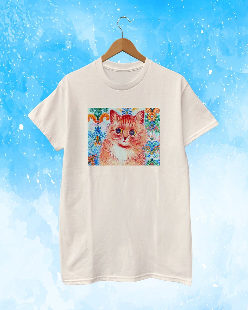 May include: A cream-colored t-shirt featuring a colorful cat portrait. The cat has orange fur, big blue eyes, and a white chest. The background is a decorative pattern with floral and geometric elements in various colors.
