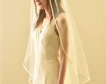 Könnte beinhalten: Ein weißer Braut Schleier mit Satin-Rand. Der Schleier wird über einem weißen Kleid getragen.