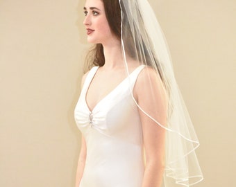 Könnte beinhalten: Eine Braut in einem weißen, ärmellosen Brautkleid mit herzförmigem Ausschnitt und einem einzigen Strassstein-Verzierungen. Sie trägt einen einlagigen Schleier in Weiß mit Satin-Saum.