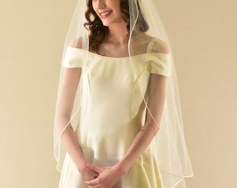 Könnte beinhalten: Eine Frau in einem weißen Brautkleid mit Schleier lächelt, während sie nach unten schaut. Das Kleid hat einen herzförmigen Ausschnitt und ein tailliertes Oberteil. Der Schleier ist schlicht und elegant.