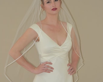 Könnte beinhalten: Eine Frau in einem weißen Brautkleid mit einem herzförmigen Ausschnitt und einem weißen Schleier mit Satinbesatz. Sie steht mit den Händen auf den Hüften.