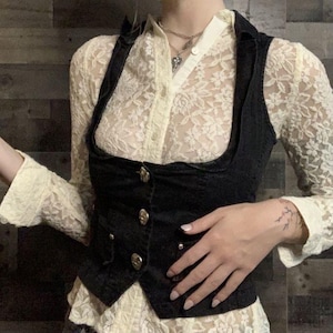 May include: A black vest with gold buttons is worn over a cream-colored lace shirt. The vest has a unique curved design and a tailored fit. The shirt has long sleeves and a delicate floral pattern. A silver necklace is visible.