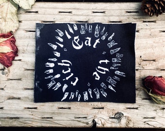 May include: A black patch with white text and teeth illustrations. The text reads "Eat the Rich" in a circular arrangement. The patch is on a wooden surface with dried roses, leaves, and a pine cone.