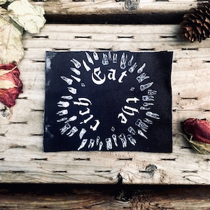 May include: A black patch with white text and teeth illustrations. The text reads "Eat the Rich" in a circular arrangement. The patch is on a wooden surface with dried roses, leaves, and a pine cone.