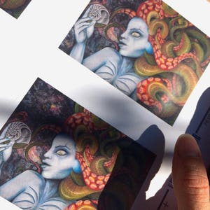 Puede incluir: Impresión artística que representa una figura de piel pálida con detalles azules y cabello con forma de tentáculos en tonos naranja y verde. La figura sostiene una concha de nautilus. La obra tiene un tema de fantasía y está impresa en papel cuadrado.