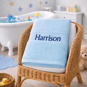 May include: A light blue bath towel featuring the name Harrison embroidered in dark blue thread. The towel is folded and draped over a woven wicker chair. The fabric has a soft, textured terry cloth finish with a smooth band across the centre.