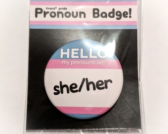 Può includere: Un badge rotondo per i pronomi con un bordo a strisce blu, rosa e bianco. Il badge dice "HELLO my pronouns are she/her" in testo nero.