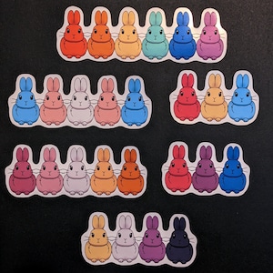Puede incluir: Una colección de pegatinas de conejos coloridos en varios tamaños. Cada pegatina presenta una fila de tres a cinco conejos de dibujos animados en tonos de rojo, naranja, amarillo, verde, azul, morado y blanco. Las pegatinas están dispuestas sobre una superficie negra.