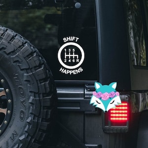 May include: Black Jeep with a white sticker that says "SHIFT HAPPENS" with a gear shift pattern inside a circle. There is a small fox sticker with pink flowers on its head on the back of the Jeep.