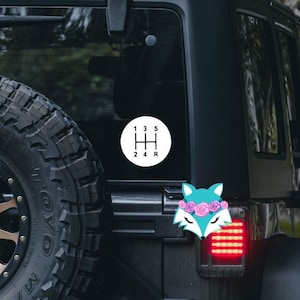 Può includere: Una Jeep nera con un adesivo bianco di uno schema di cambio marcia sul lunotto posteriore. C'è anche un adesivo di una volpe rosa e blu con una corona di fiori sul retro della Jeep.