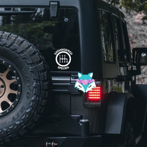 May include: A black Jeep with a white sticker on the back window that says "Endangered Species" with a gear shift pattern inside a circle. There is a pink fox sticker with flowers on its head on the back of the Jeep.