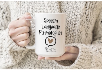 Op de afbeelding: Een witte keramische koffiemok met zwarte tekst die "Speech Language Pathologist" luidt met een spraakballon die een hart met een luipaardprint bevat. De naam "Katie" staat onder de spraakballon.