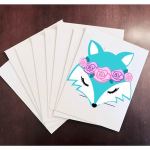 May include: A stack of white cardstock paper with a turquoise fox face with closed eyes and a pink and purple flower crown on the top sheet.