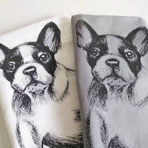 May include: Two tea towels with a black and white illustration of a French Bulldog. One towel is white with a black design, and the other is gray with a black design.