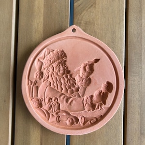 May include: A circular, terracotta-coloured Christmas ornament with a relief of Santa Claus, pointing at a toy car and an owl. The ornament has a hole at the top for hanging, and is made of a reddish-brown material.