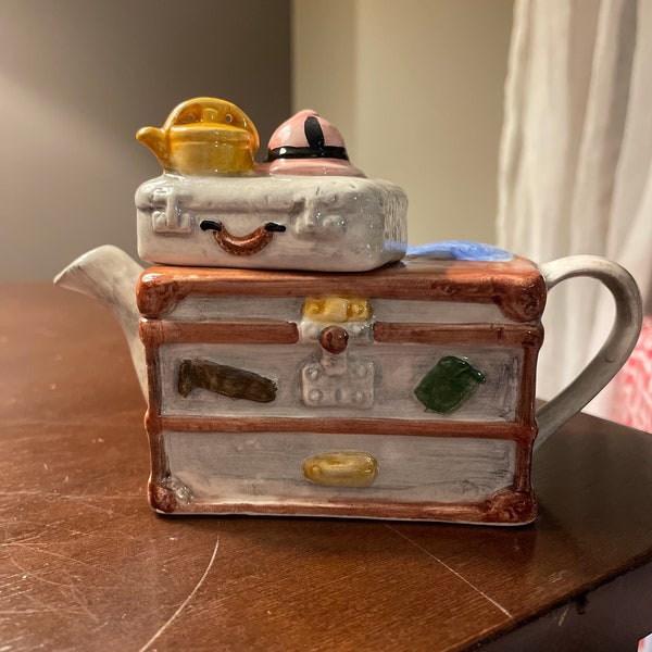 Vintage Decorative Ceramic Teapot Dresser With Luggage, Pink Hat, and Yellow Kettle.