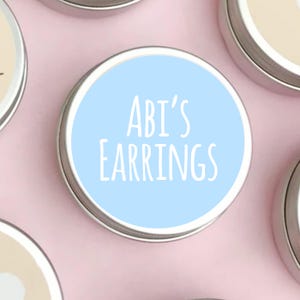 May include: A circular silver tin with a pale blue lid. The lid is printed with the white text "ABI'S EARRINGS". The tin is likely used for storing or packaging earrings, presented on a pink surface.