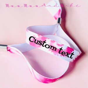 May include: Pink and white tie-dye fabric wristband with a white elastic band and a silver metal clasp. The wristband has black text that says "Custom text".