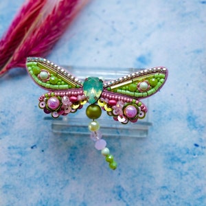 May include: A green and pink beaded dragonfly brooch with a clear crystal center. The brooch is on a light blue background.
