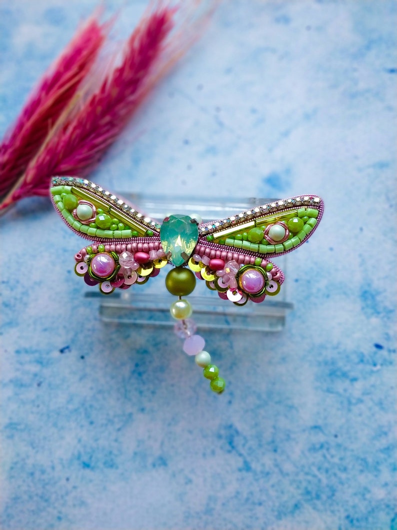 May include: A green and pink beaded dragonfly brooch with a clear crystal center. The brooch is on a light blue background.