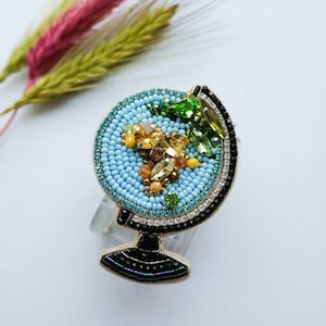 K&ouml;nnte beinhalten: Eine Perlen-Globus-Brosche mit blauem Hintergrund, goldenen Akzenten und gr&uuml;nen und orangefarbenen Edelsteinen. Die Brosche befindet sich auf einem wei&szlig;en Hintergrund.