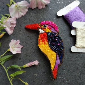 May include: A colorful beaded bird brooch with a pink, purple, yellow, orange, and blue design. The bird has a long, thin beak and a round body. The brooch is handmade and would make a unique gift.